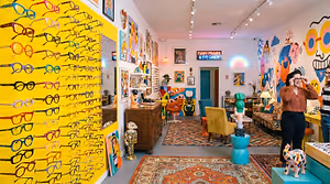 An interior photo of a store called Funky Frames, showcasing colorful paintings on the walls, a table with eyeglasses on display, and a couch with a pillow.