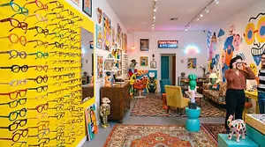 An interior photo of a store called Funky Frames, showcasing colorful paintings on the walls, a table with eyeglasses on display, and a couch with a pillow.