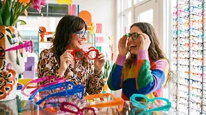 Two women are wearing colorful glasses and smiling in a shop with many colorful glasses on display.
