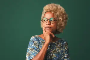 A woman with curly blonde hair wearing glasses is posing for a photo with her hand on her chin.