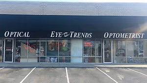 A storefront for Eye & Trends Optometrist, featuring a black awning with white lettering, a large glass window displaying various items, and a parking lot in front.