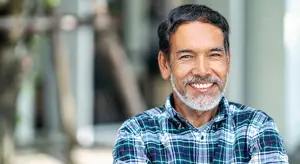 A man with a beard and mustache is smiling and posing for a photo outside a building with a glass wall and a tree in the background