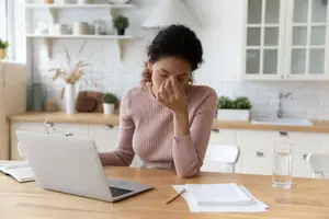 A woman sitting at a desk, holding her nose and covering her mouth with her hand, probably feeling sick. There is a laptop, a glass, a book, and a pen on the desk. There is a cabinet with a potted plant, a vase, and a glass in the background. There is a sink with a faucet in the right corner.