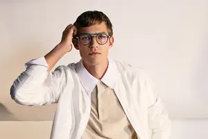 A man in a white coat is adjusting his glasses in a studio setting.