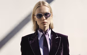 A woman with blonde hair wearing a purple velvet suit and glasses standing against a white wall