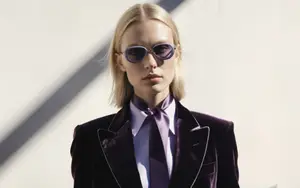 A woman with blonde hair wearing a purple velvet suit and glasses standing against a white wall