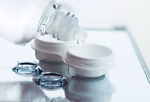 A close-up of a person holding a bottle of eye drops and pouring it into a lens case on a reflective surface.