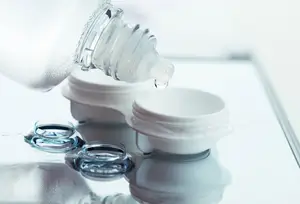 A close-up of a person holding a bottle of eye drops and pouring it into a lens case on a reflective surface.