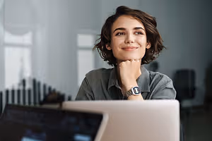 A woman is smiling and looking at the camera while sitting in front of a laptop
