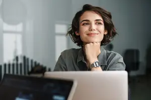 A woman is smiling and looking at the camera while sitting in front of a laptop