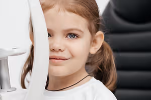 A young girl with brown hair wearing a white shirt and a black necklace is having her eyes examined by an optometrist using a white ophthalmoscope with a silver handle.