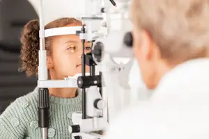A little girl is looking at the camera with her eyes closed while wearing a green sweater and sitting in front of an eye exam machine in a blurry background with a doctor standing next to her