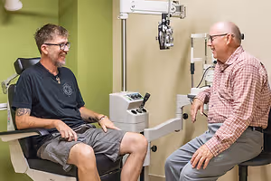 An eye doctor is sitting in a chair while talking to a patient sitting in another chair next to him with an eye machine in the background.