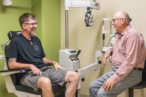 An eye doctor is sitting in a chair while talking to a patient sitting in another chair next to him with an eye machine in the background.