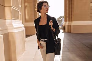 A woman wearing glasses and a black blazer stands on a sidewalk in front of a building.