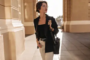 A woman wearing glasses and a black blazer stands on a sidewalk in front of a building.