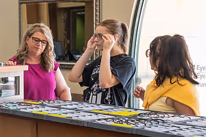Three women, one wearing glasses, are standing behind a desk with a variety of glasses on display. They are likely at an optical shop, with a mirror and window behind them.