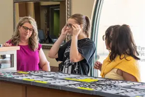 Three women, one wearing glasses, are standing behind a desk with a variety of glasses on display. They are likely at an optical shop, with a mirror and window behind them.