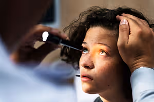 A woman in a medical setting is having her eyes examined by a doctor with a flashlight