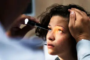 A woman in a medical setting is having her eyes examined by a doctor with a flashlight