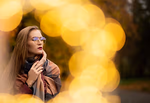 A woman wearing glasses and a scarf is standing and posing for a photograph in a park with a blurry background of yellow lights.