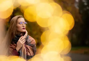 A woman wearing glasses and a scarf is standing and posing for a photograph in a park with a blurry background of yellow lights.