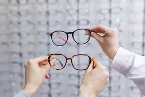 Optometrist holding two pairs of glasses in front of a wall with several pairs of glasses displayed on the wall behind them