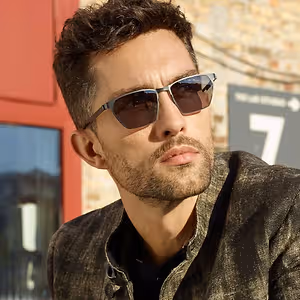 A man with curly hair and beard wearing sunglasses and a brown jacket standing in front of a brick wall