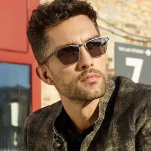 A man with curly hair and beard wearing sunglasses and a brown jacket standing in front of a brick wall
