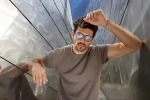 A man in a brown t-shirt and sunglasses standing near a reflective wall.