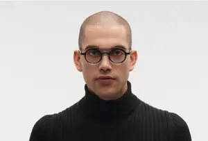 A bald man wearing a black turtleneck and glasses with a serious expression