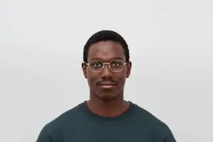 A man with glasses and a green shirt standing and smiling in front of a white background.