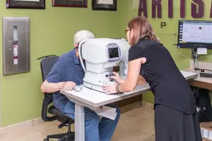 An older man is having his eye examined by a woman with a white machine on a white table in a room with green walls and a computer monitor