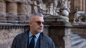 A man in a business suit stands in front of a building with a stone lion statue.