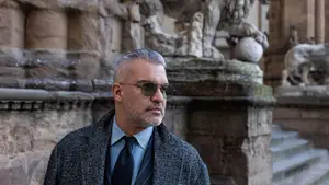 A man in a business suit stands in front of a building with a stone lion statue.