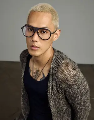 A person wearing a black shirt, glasses, and a necklace with a gray sweater on a couch.