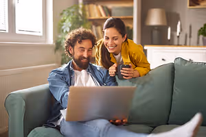 A man and a woman are sitting on a couch and looking at a laptop. The man is holding a cup and the woman is smiling.