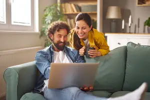 A man and a woman are sitting on a couch and looking at a laptop. The man is holding a cup and the woman is smiling.