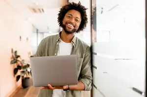 A smiling man in a green shirt and white undershirt is holding a laptop in a hallway next to a plant and glass wall.