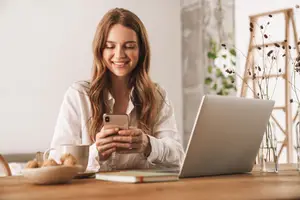 A smiling woman using a cell phone while sitting at a desk with a laptop and cup
