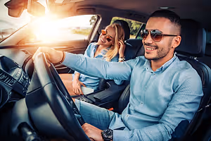 A man and woman sit inside a car with the woman smiling and the man holding the steering wheel. The car is parked on a sunny day.