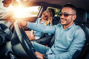 A man and woman sit inside a car with the woman smiling and the man holding the steering wheel. The car is parked on a sunny day.