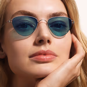 A close-up of a woman with blonde hair wearing silver-framed sunglasses with green lenses, touching her face.