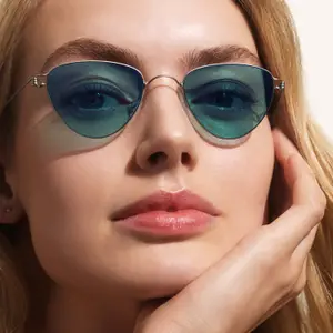 A close-up of a woman with blonde hair wearing silver-framed sunglasses with green lenses, touching her face.