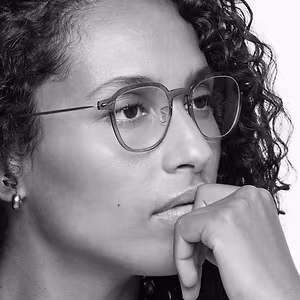 A black and white portrait of a woman with curly hair wearing glasses and a small earring in her left ear, she is biting her bottom lip and has her left hand to her chin.