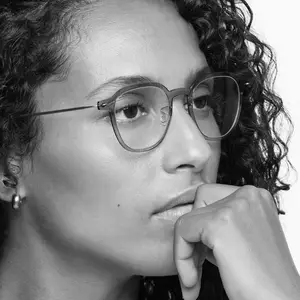 A black and white portrait of a woman with curly hair wearing glasses and a small earring in her left ear, she is biting her bottom lip and has her left hand to her chin.