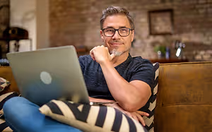 A man is sitting on a couch and looking at a laptop