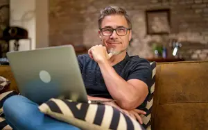 A man is sitting on a couch and looking at a laptop