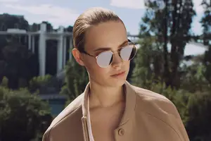 A woman with short hair wearing sunglasses and a beige jacket is standing in front of a bridge and trees