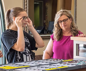 A woman is trying on glasses in front of a woman with blonde hair in a pink shirt at an optical shop